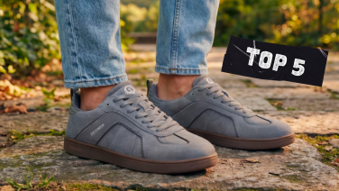 A close-up of both feet in light grey suede trainers with brown rubber soles and a white Giesswein logo, on cobblestones bathed in autumnal light.