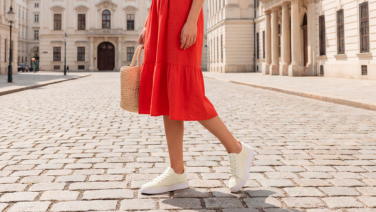 A woman in a red summer dress, carrying a wicker bag, is walking down a cobbled alley in the old town, wearing white knitted trainers.