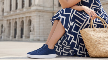 A woman wearing a blue and white patterned summer dress is sitting on a step in front of a historic town building; she is wearing navy blue knitted trainers and is holding the handle of a woven basket bag.