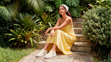 A woman is sitting on a stone step in a lush garden. She is wearing a yellow summer dress, a white hairband and cream-coloured Giesswein trainers.