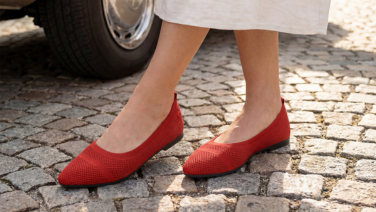 A close-up of feet in red Giesswein knitted ballerina flats, standing on cobblestones next to a parked car.
