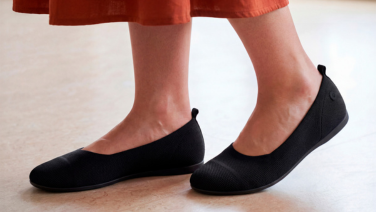 A close-up of feet wearing black Giesswein knitted ballerina flats with a pull tab at the heel, beneath a terracotta-coloured skirt, on a light-coloured floor.