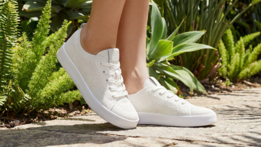 A close-up of feet in white Giesswein knitted trainers with white soles, standing on a stone path amongst green plants.