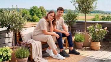 Two people are sitting on a wooden bench on a terrace with lavender and potted plants; they are wearing white and black Giesswein Merino Runner trainers.