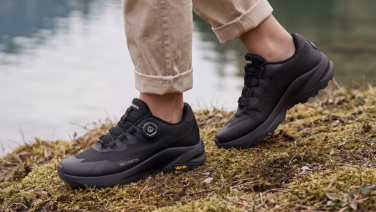 A close-up of feet in black Giesswein trainers with a BOA twist-lock fastening and Vibram soles, standing on a moss-covered riverbank.