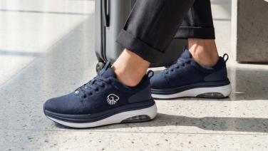 A close-up of a person’s feet resting on a terrazzo floor. They are wearing dark blue merino wool trainers, featuring a white sole and a visible Air-Cushion element at the heel. The Giesswein logo is visible on the side of the shoe.