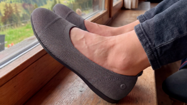 A close-up of a person’s feet, resting on a wooden window sill with their dark trousers rolled up. They are wearing taupe-coloured, textured knitted ballet flats with dark soles. A green mountain landscape can be seen through the window.