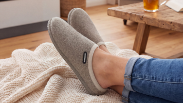 A close-up of a person’s feet, crossed over one another, wearing jeans and lying on a cream-coloured knitted blanket; in the background is a wooden table with a drink and a book. She is wearing grey merino wool slippers with a light-coloured trim.