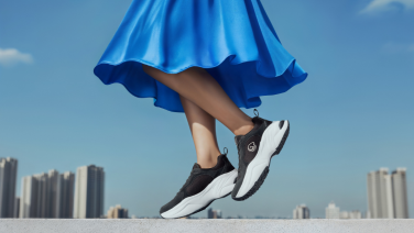 A woman in a cobalt-blue dress is jumping lightly on the roof of a high-rise building, wearing black Giesswein trainers with white soles, against a city skyline in the background.