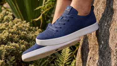 A close-up of a dark blue Giesswein Merino trainer with a white flat sole, set against a backdrop of Mediterranean plants and stones.