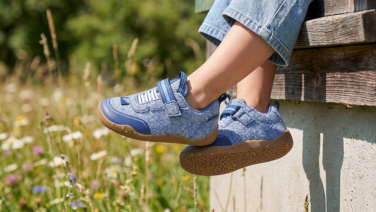 Close-up of a light-blue pair of Giesswein kids’ trainers with hook-and-loop fastener and a honey-coloured sole, worn with jeans in front of a blooming summer meadow.