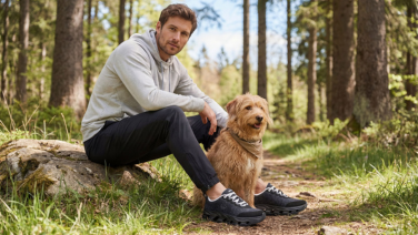 A man wearing a grey hoodie and dark trousers is sitting on a tree trunk in the woods next to a Goldendoodle, wearing black Giesswein outdoor trainers.