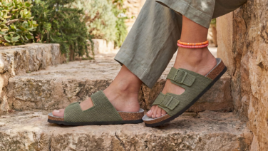 Feet in olive-green knitted sandals with two buckles and a cork insole, standing on Mediterranean stone steps, paired with olive-green linen trousers and colourful anklets.