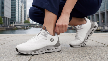 A person is crouching on a paved path against a city backdrop, wearing white Giesswein Wool Boom trainers with a textured outsole.