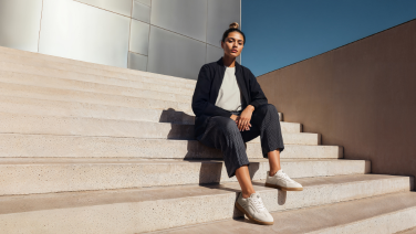 A woman wearing a dark blue blazer and grey trousers is sitting on stone steps in front of a modern building, wearing white Giesswein merino trainers.