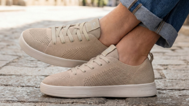 A close-up of a sand-coloured Giesswein trainer with a textured knitted upper and a white sole. The person is wearing light-coloured jeans and is sitting on cobblestones.