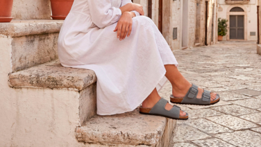 A woman in a white dress is sitting on a stone staircase in a Mediterranean alleyway; she is wearing grey Giesswein sandals with two straps and a cork insole.