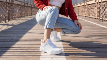 A close-up of the feet of a person wearing a red jacket and light-coloured jeans, sitting in a squatting position on a wooden bridge with a white marking line. They are wearing all-white, sporty trainers.