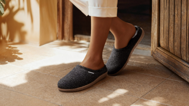 Feet in white trousers and dark grey woollen slippers on light-coloured tiles, warm sunlight falling on the floor.