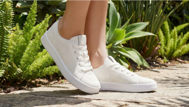 A close-up of feet in white Giesswein knitted trainers with white soles, standing on a stone path amongst green ferns.