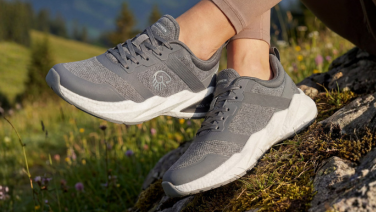 A close-up of a person’s feet standing on a moss-covered rock in a mountainous landscape. They are wearing grey, sporty trainers with white outsoles.