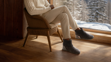 Person in beige casual wear with dark grey Giesswein woollen slippers sits relaxed by the window overlooking a snowy winter landscape.