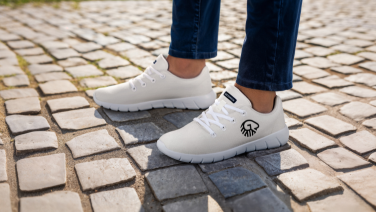 Close-up of white Giesswein merino sneakers with a black brand logo on the feet of a person in jeans, standing on cobblestones.