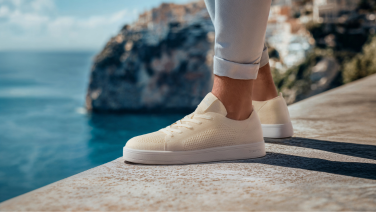 Close-up of cream-coloured Giesswein Merino sneaker with white flat sole on a stone wall in front of a Mediterranean sea backdrop.