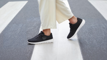 Close-up of black Giesswein Merino sneakers with a white sole on the feet of a person wearing wide white trousers on a zebra crossing.