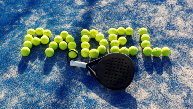 A black padel racket lies on a blue hard court. Yellow-green tennis balls are arranged next to it to spell out the word “PADEL”.