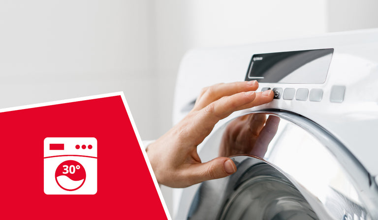 A person's hand adjusting the control dial of a white washing machine. A red icon shows a washing machine symbol with "30°" indicating machine washable at 30 degrees Celsius.