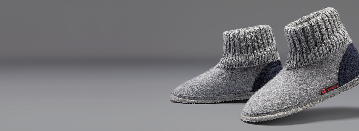 A pair of gray wool slipper boots with ribbed knit ankle cuffs and navy blue heel accents, featuring the Giesswein brand label, against a neutral gray background.