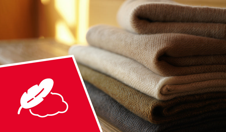 A stack of neatly folded beige and brown knitted wool sweaters or garments sits on a wooden surface in warm, natural lighting. In the foreground is a red square icon featuring a white symbol of a leaf crossed out over a cloud shape, indicating hypoallergenic or allergen-free properties. The soft, textured fabrics appear cozy and luxurious in the golden ambient light.
