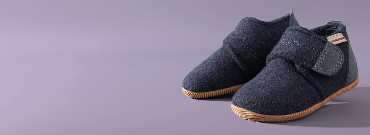 A pair of navy blue children's Giesswein shoes with velcro straps and tan rubber soles, made from wool felt material, against a purple background.