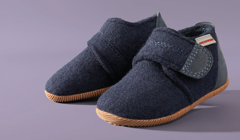 A pair of navy blue children's Giesswein shoes with velcro straps and tan rubber soles, made from wool felt material, against a purple background.