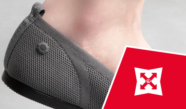 Close-up of a person's foot wearing a gray knitted slip-on shoe with textured upper and a small logo button. A red icon shows a white shield with arrows, indicating durability or strength properties.