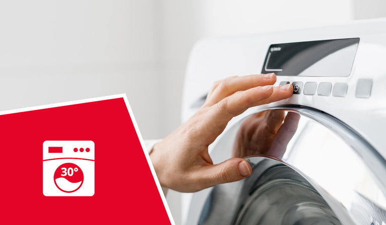 A person's hand adjusting the control dial of a white washing machine. A red icon shows a washing machine symbol with "30°" indicating machine washable at 30 degrees Celsius.