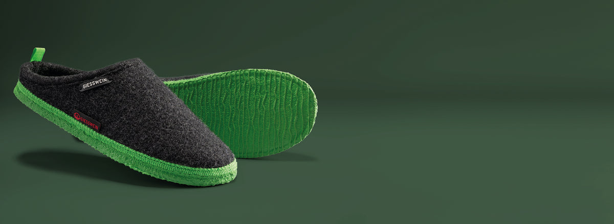 A pair of Giesswein slippers with dark gray wool uppers and bright green textured soles, shown from side and bottom views against a green background.