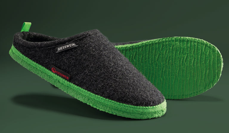 A pair of Giesswein slippers with dark gray wool uppers and bright green textured soles, shown from side and bottom views against a green background.