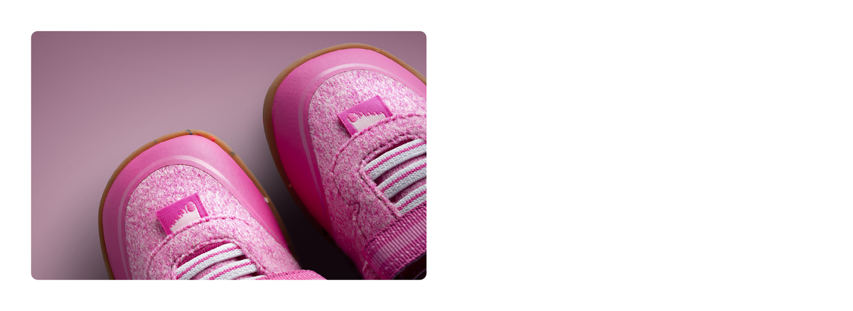 A close-up of two pink Giesswein children’s shoes with a merino upper, elastic laces and a honey-coloured sole against a pink background.