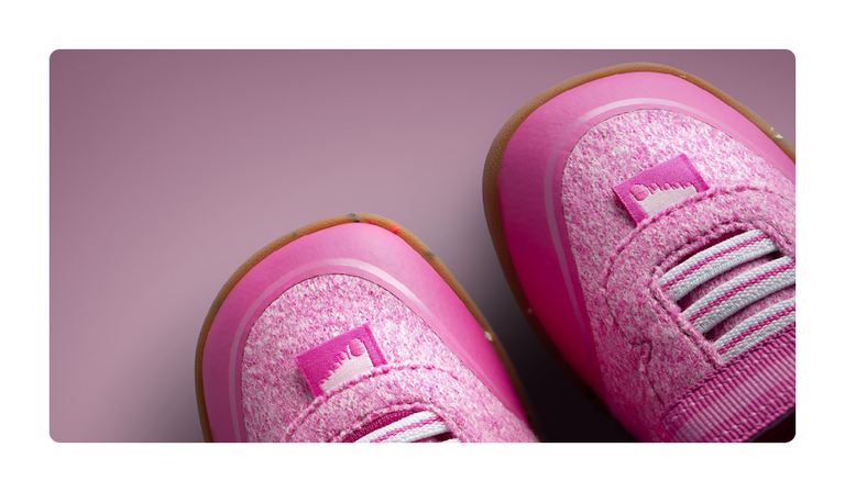 A close-up of two pink Giesswein children’s shoes with a merino upper, elastic laces and a honey-coloured sole against a pink background.